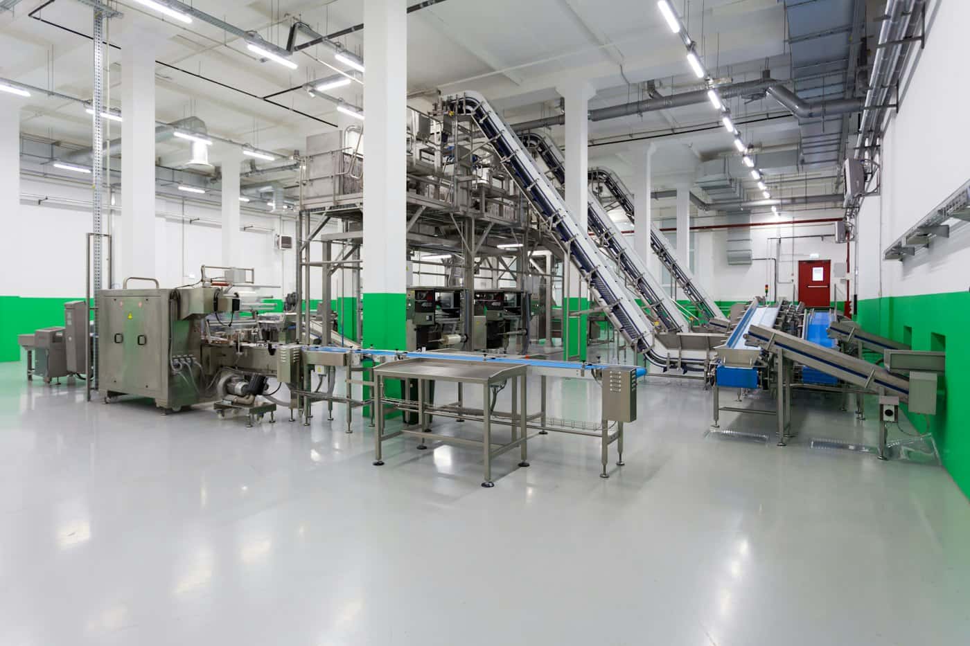 Bright packaging line with stainless steel machines and inclined conveyors on a pale epoxy floor, framed by green and white walls
