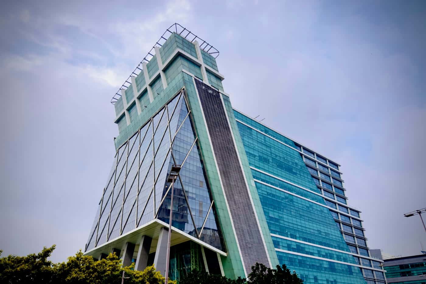 Slanted glass office tower in DLF Cyber City, Gurugram, against a cloudy sky, with trees along its base