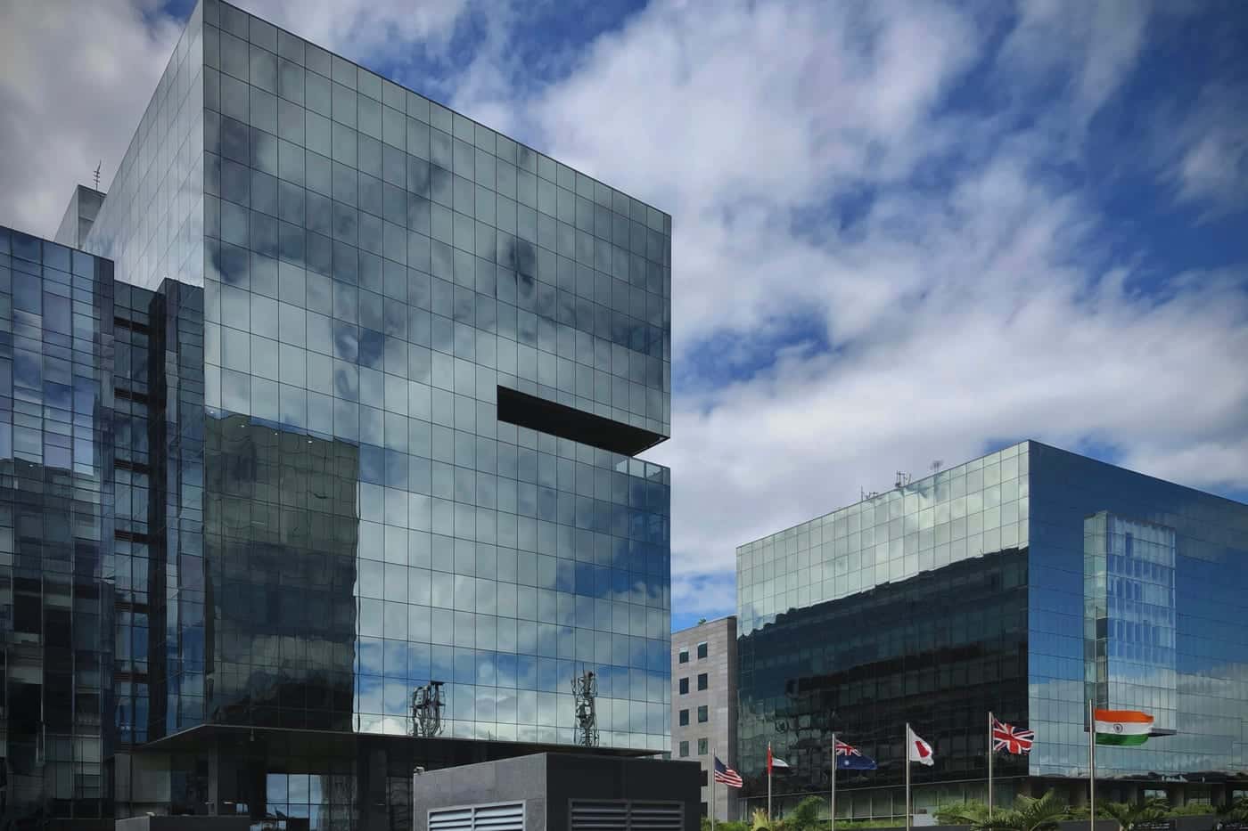 Glass office blocks of the World Trade Center in Kharadi, Pune, reflecting white clouds, with a row of flags at the entrance