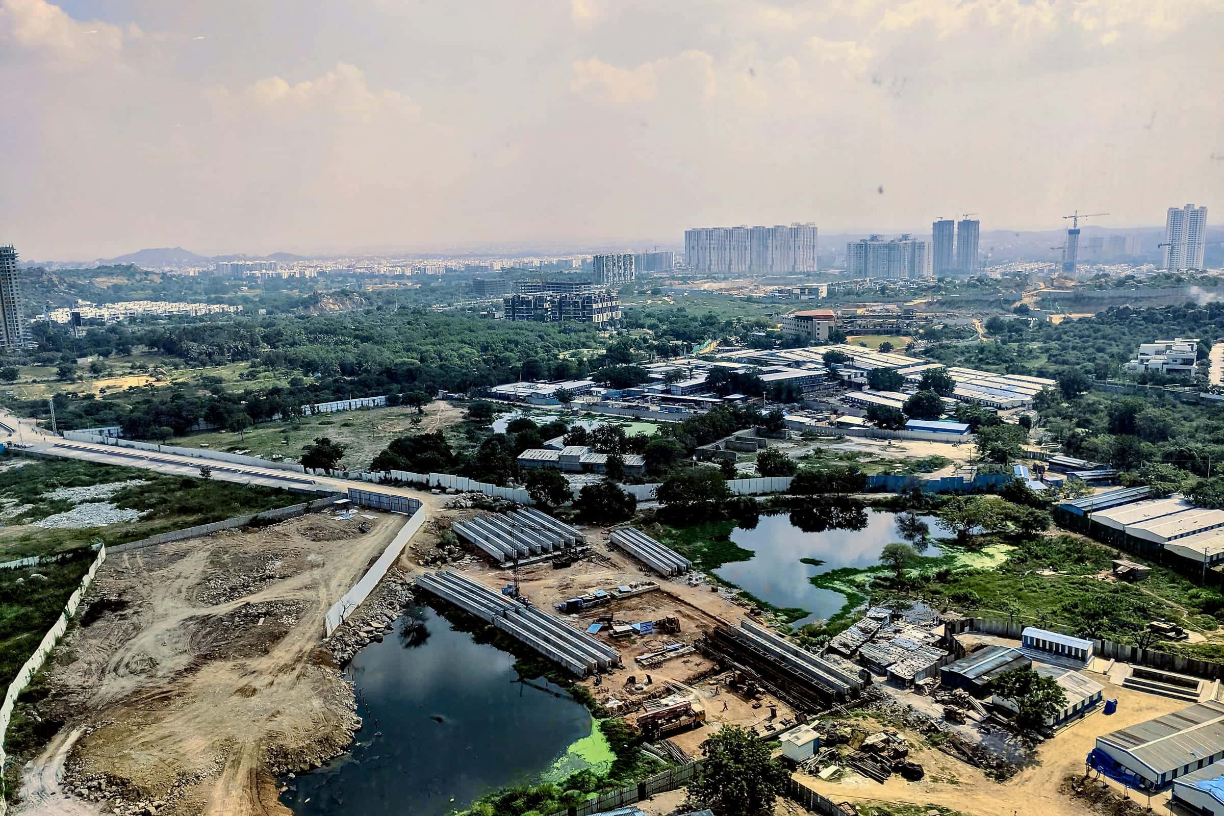 High view over cleared plots, construction yards and ponds among green scrub, with high-rise towers on the horizon