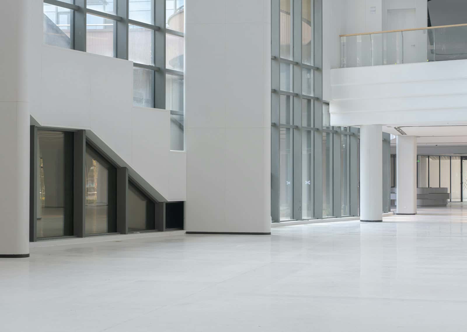 Empty white double-height hall with tall glazing, a stepped stair wall and a glass-railed mezzanine above