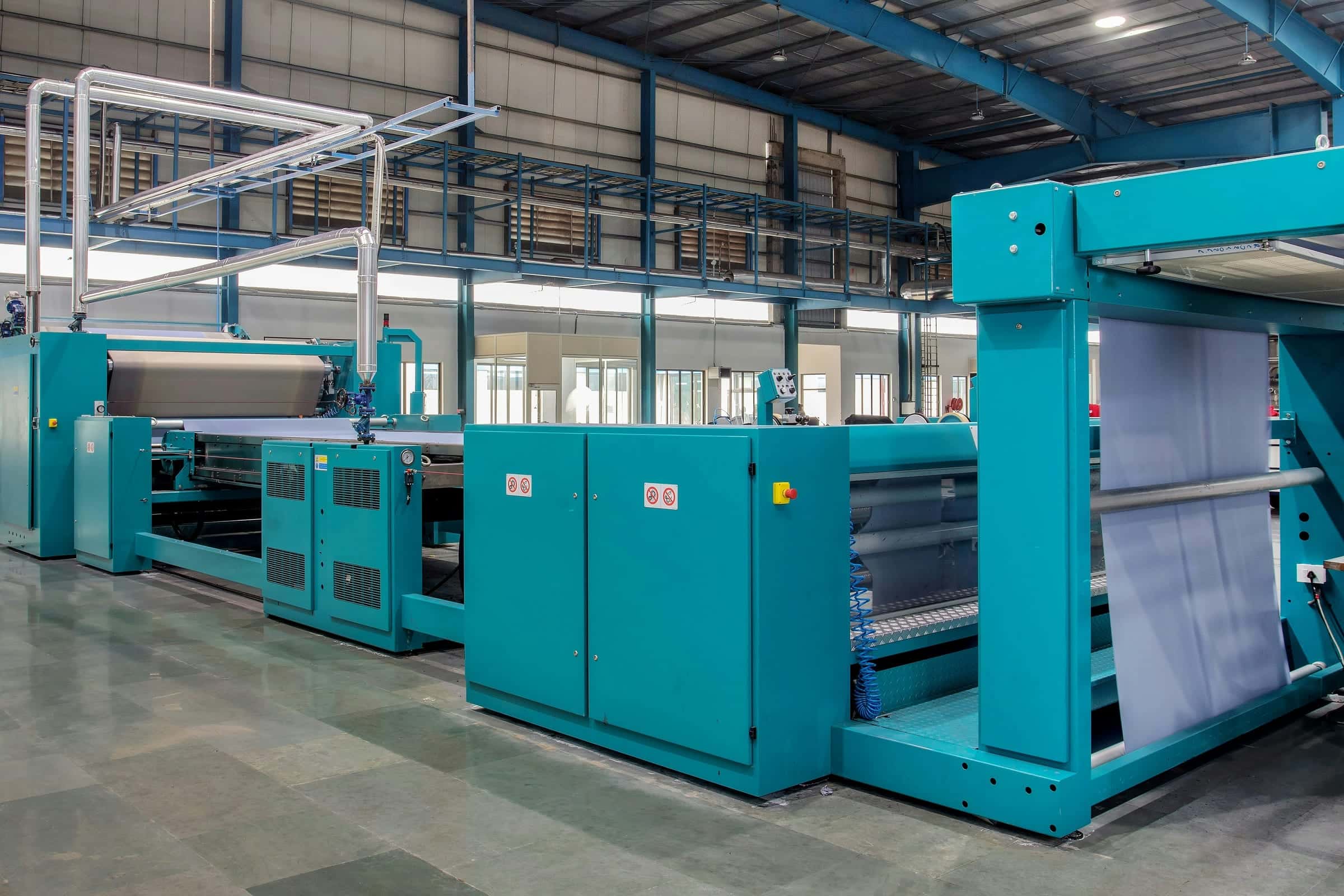 Tall steel-framed factory hall with a polished floor, a long line of teal textile finishing machines and roof lights