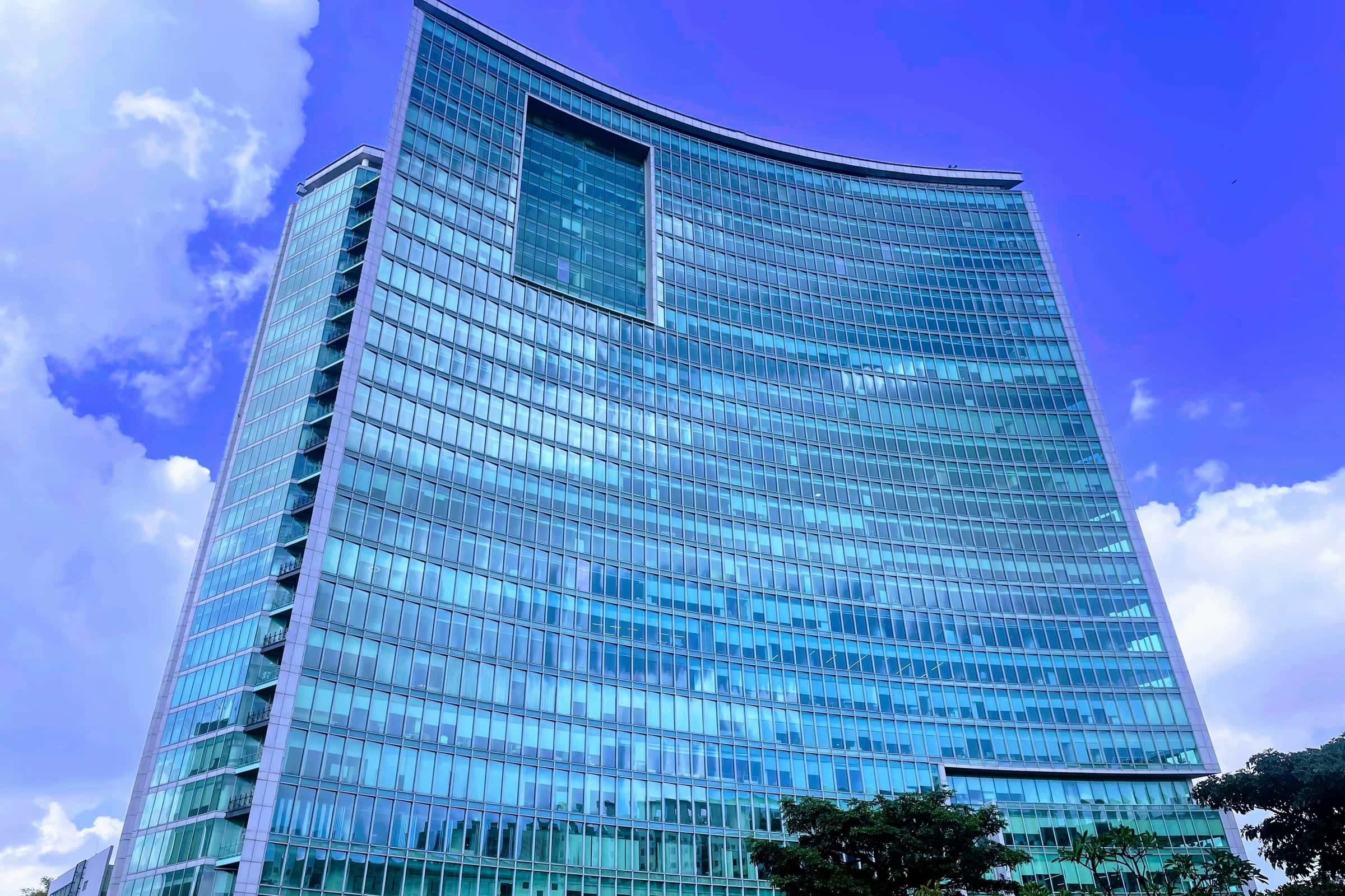 Curved glass office tower rising against a deep blue sky with white clouds, with trees at its base