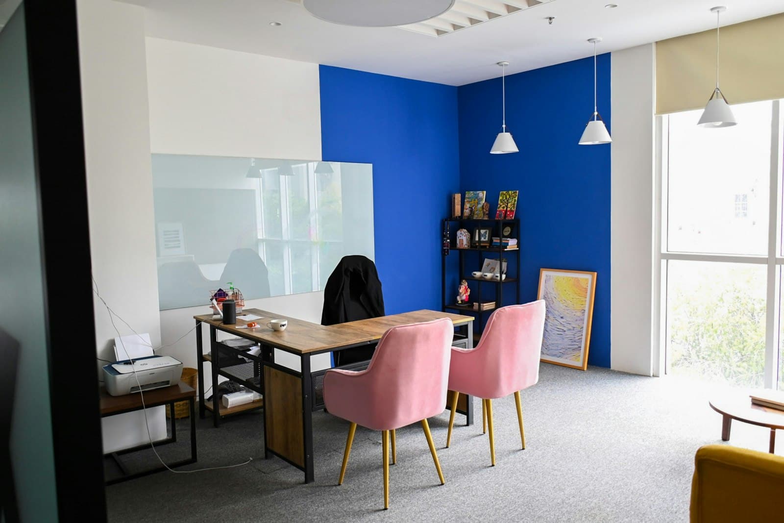Private cabin with a timber desk, two pink velvet chairs, a glass writing board and a blue feature wall by a window