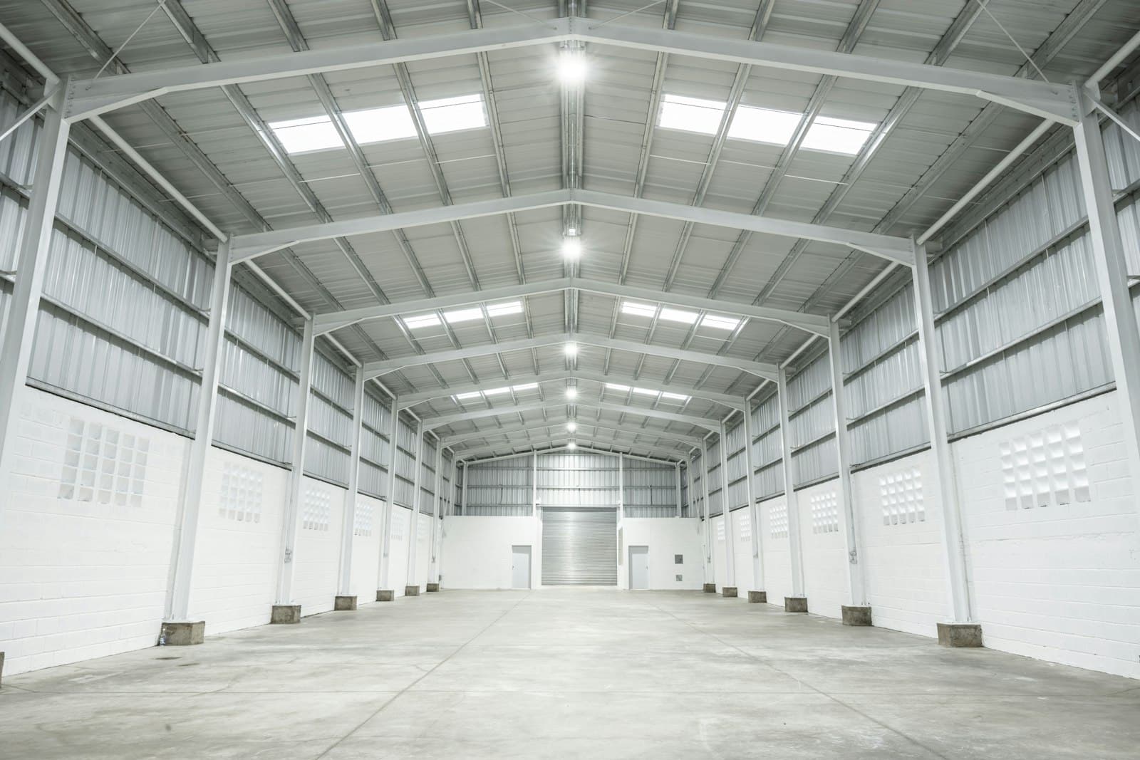 Empty warehouse hall with white steel portal frames, a high roof, skylights and a roller shutter at the far end