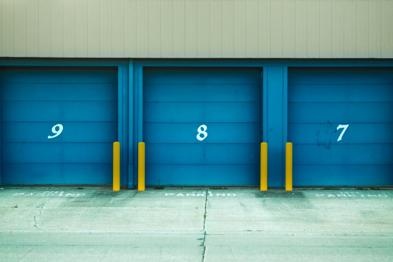 Three blue roller shutter bays numbered 9, 8 and 7 with yellow bollards, set below beige metal cladding on a concrete apron