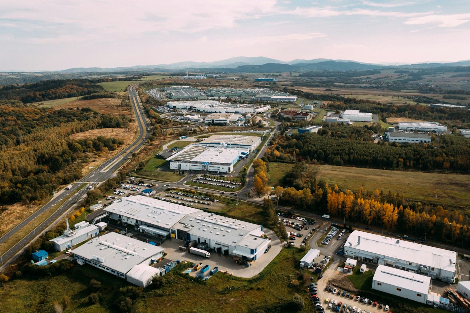Aerial view of an industrial and logistics park with large white sheds, truck yards and parking beside a highway and hills