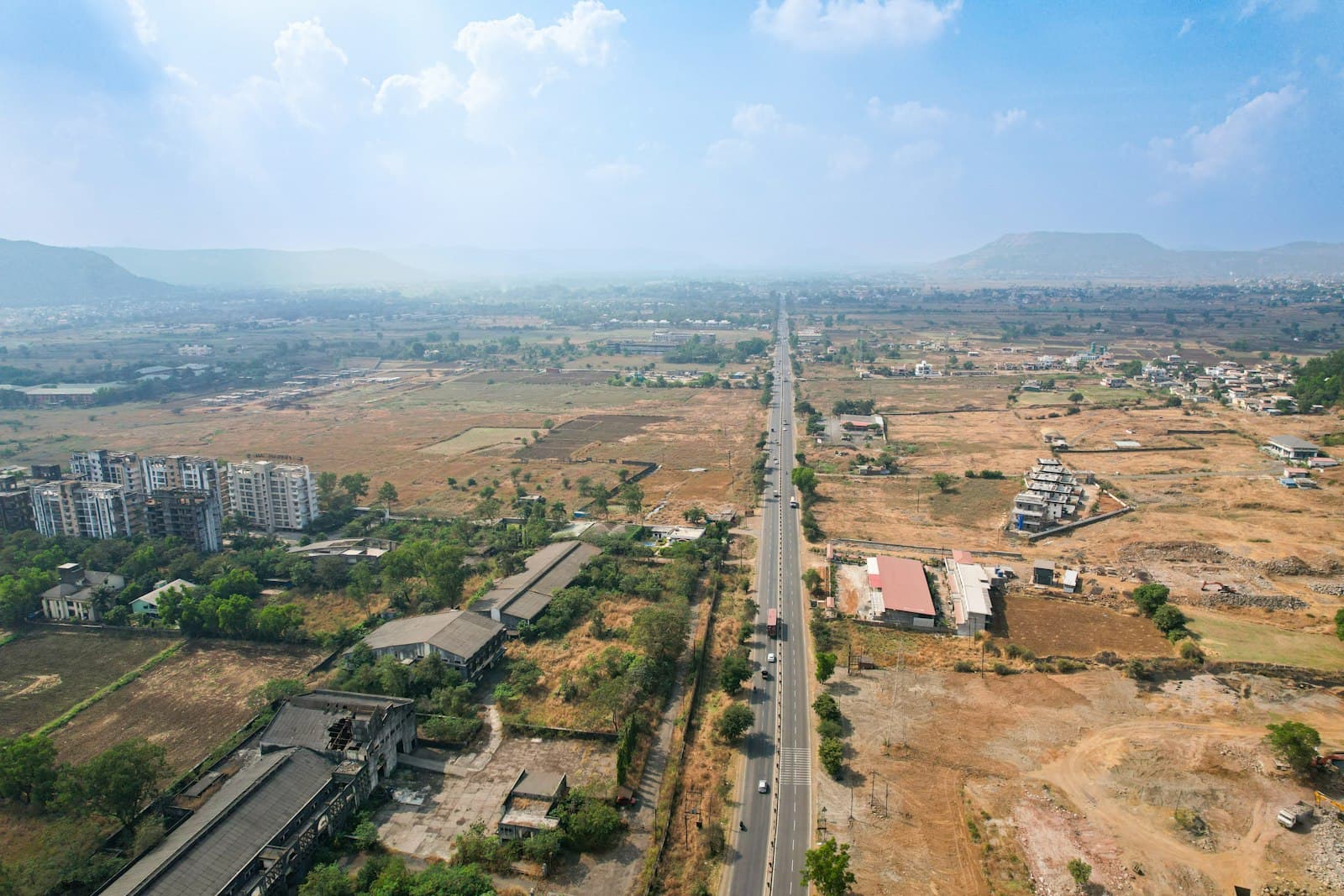 Aerial of a four-lane highway running straight past wide open plots, low buildings and distant hills