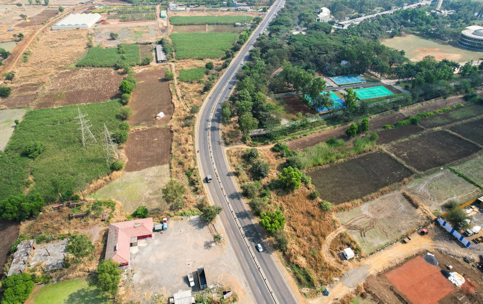 Aerial of a curving divided road flanked by cultivated and vacant land parcels, power pylons and scattered buildings