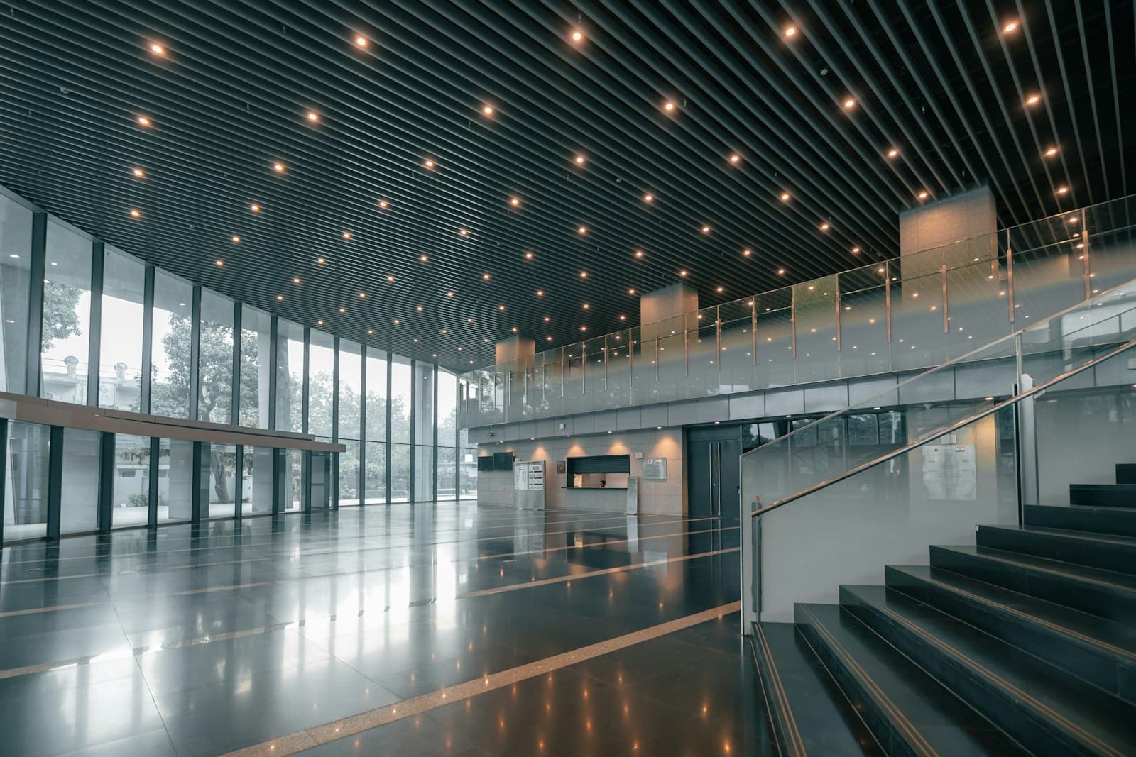 Double-height lobby with a slatted lit ceiling, glass mezzanine balcony, wide staircase and polished stone floor