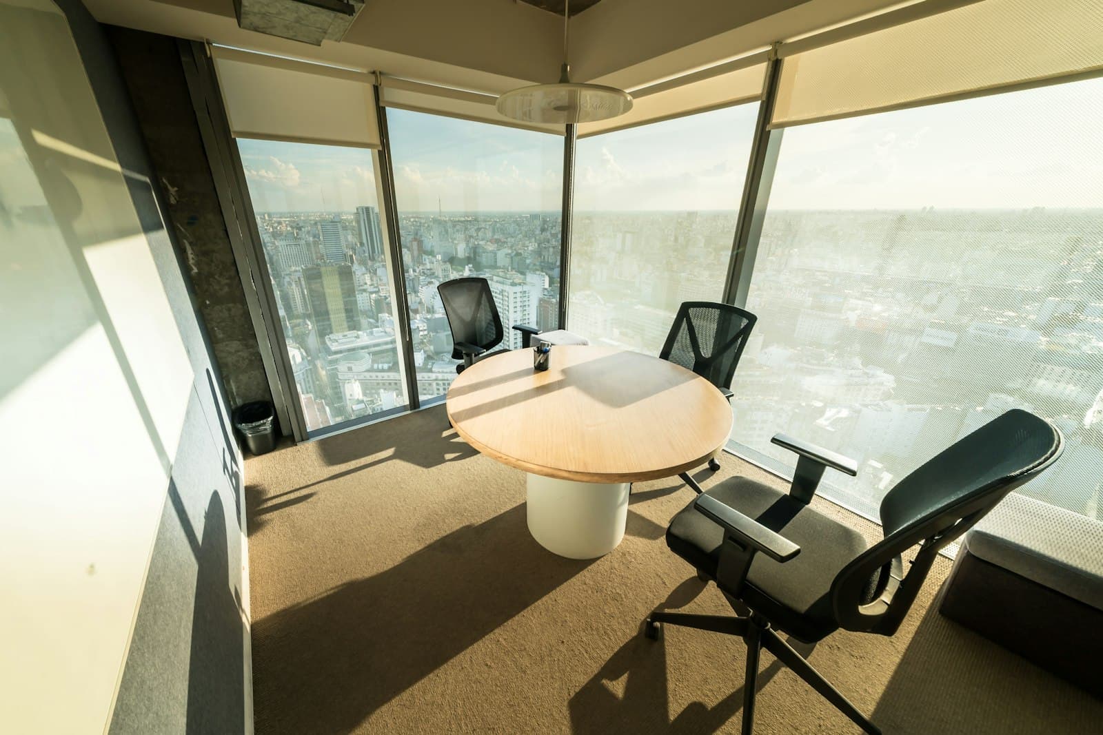 Meeting room with a round table and mesh chairs beside floor-to-ceiling windows over a sunlit city