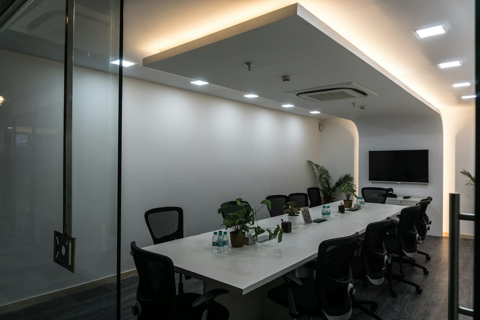 Meeting room seen through a glass wall, with a long white table, black mesh chairs, potted plants and a wall screen
