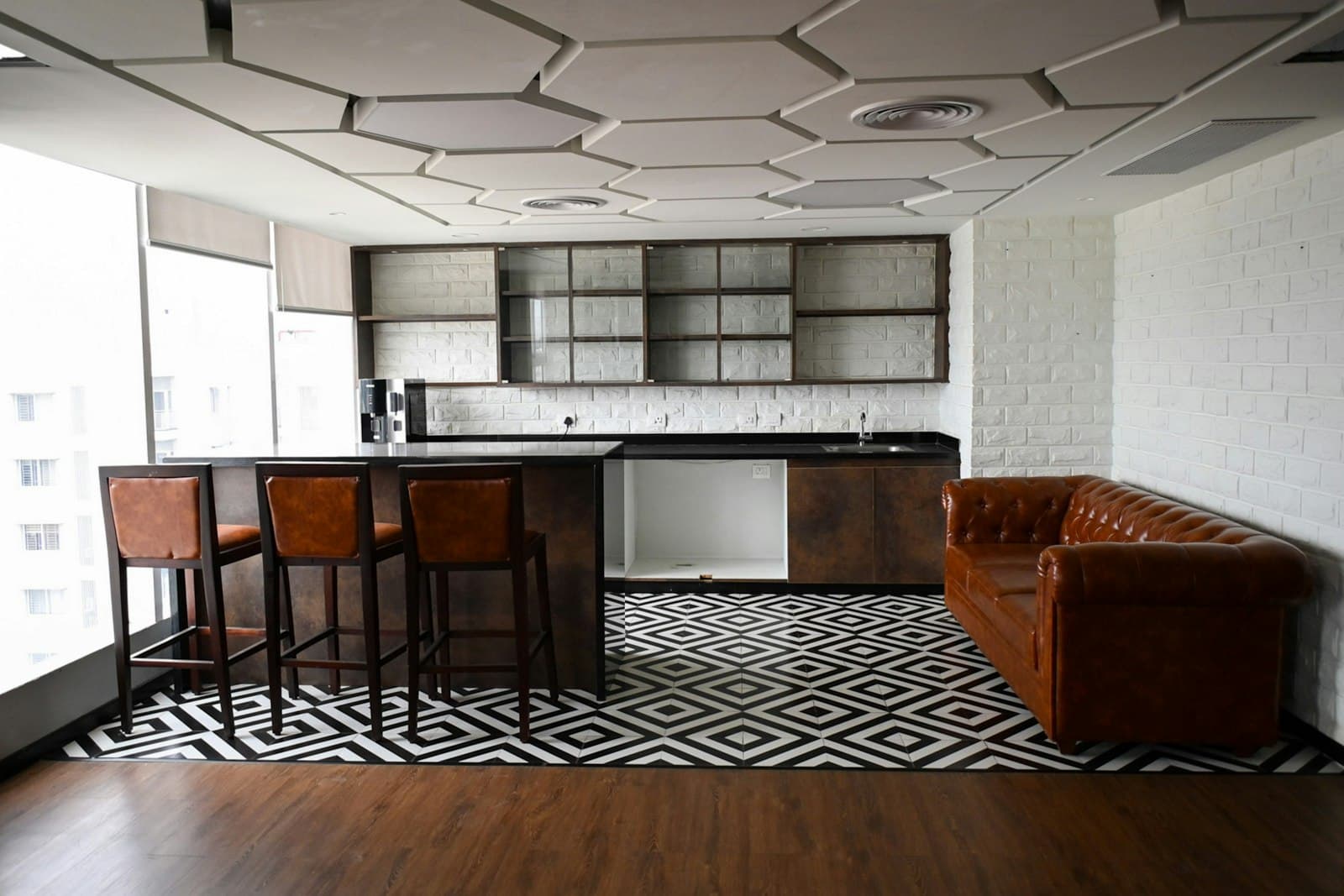 Office pantry with a dark kitchen counter, leather bar stools, a chesterfield sofa and hexagonal ceiling panels