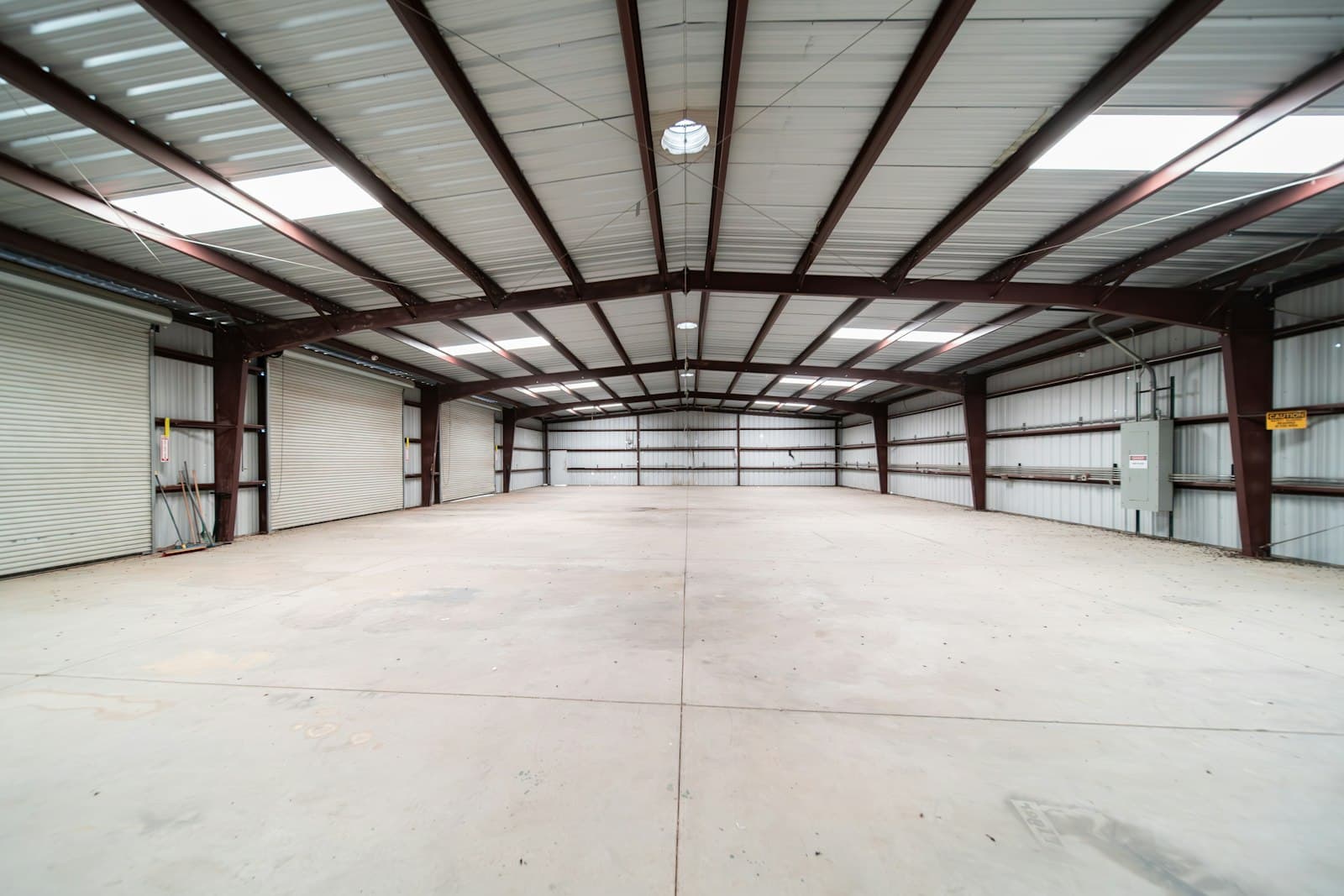 Wide empty warehouse interior with red steel roof beams, skylights, roller doors and a bare concrete floor