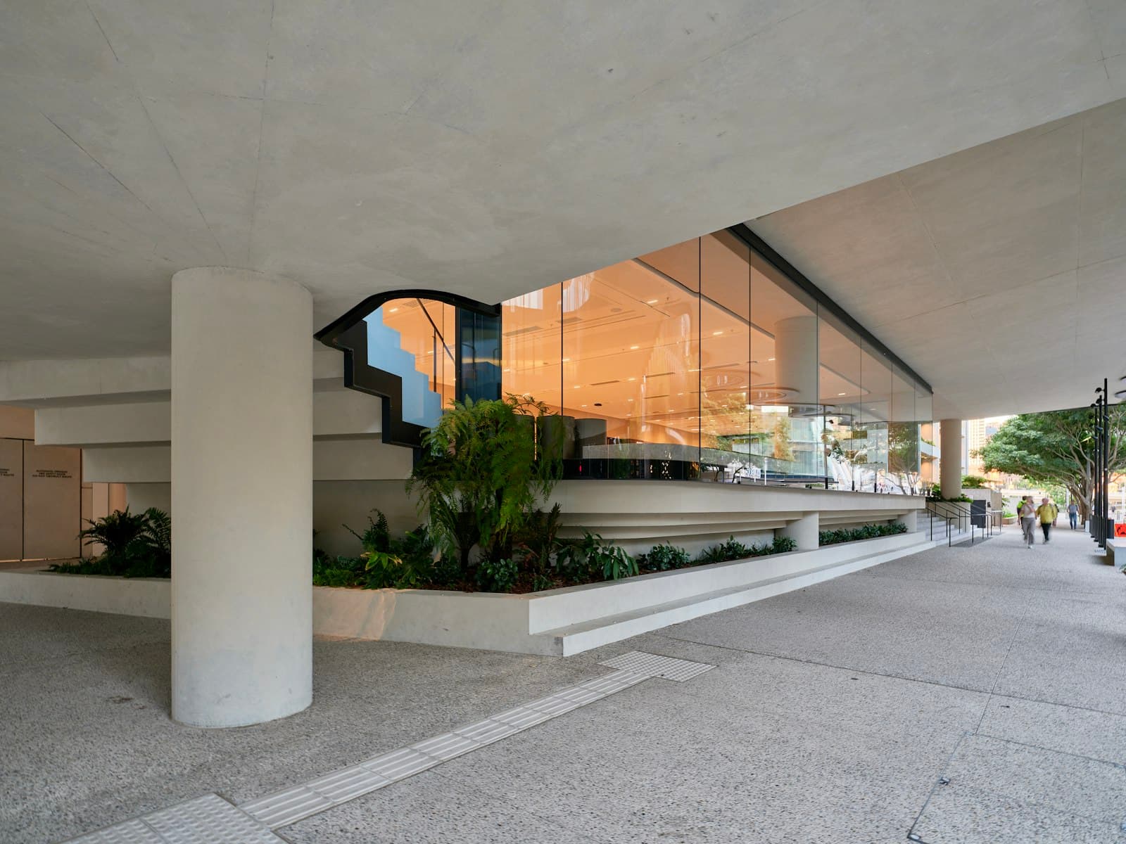 Tall glass frontage lit from within, set under a concrete canopy with tropical planting beside a wide pavement