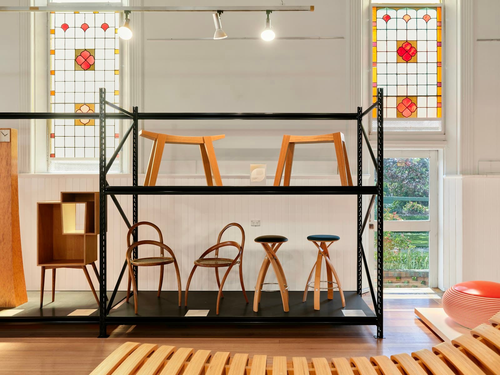 Wooden chairs and stools on black display shelving in a bright showroom with stained-glass windows and timber floor