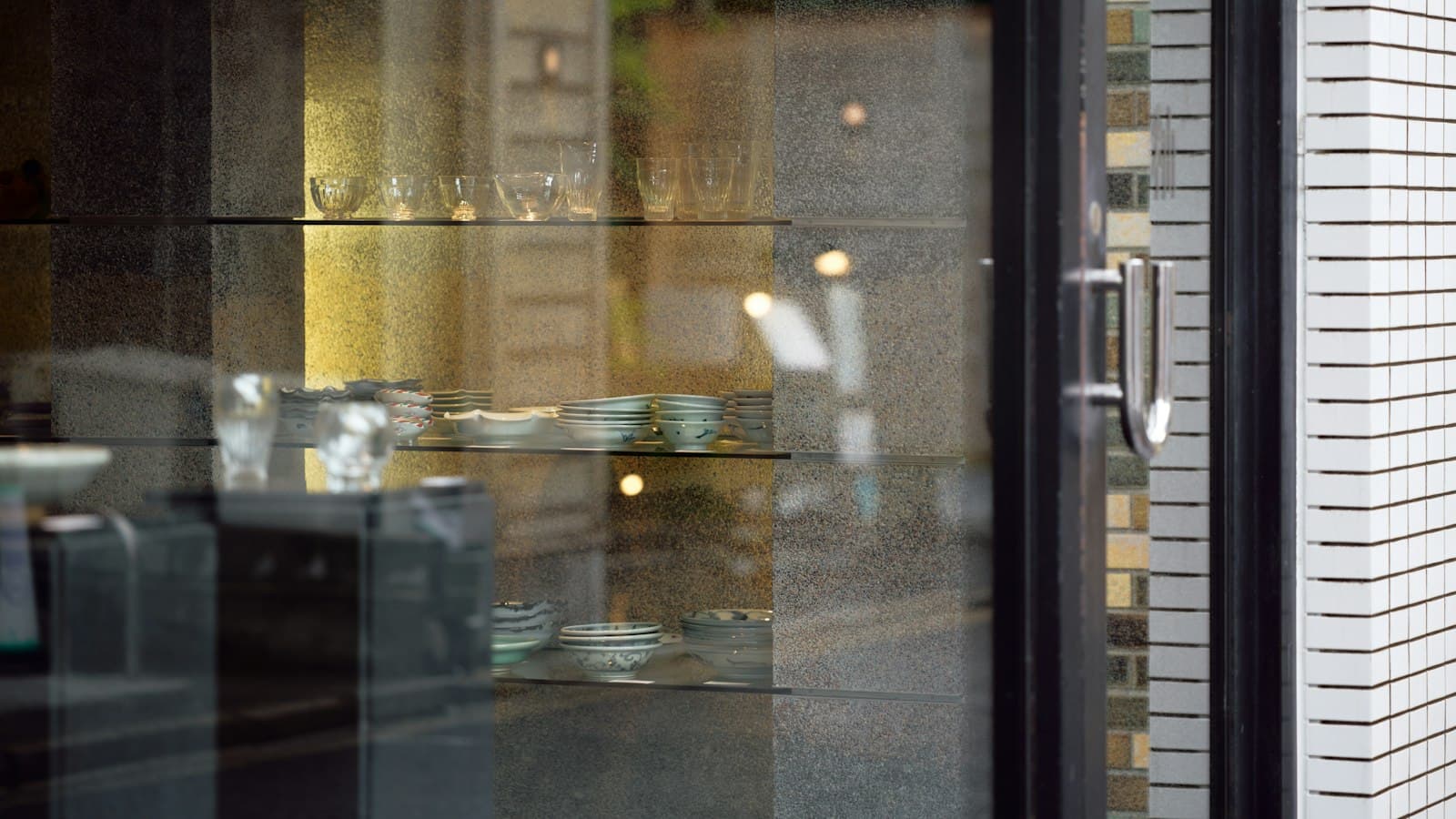 Glassware and ceramic bowls on lit glass shelves behind a shop window, beside a steel door handle and white tiles