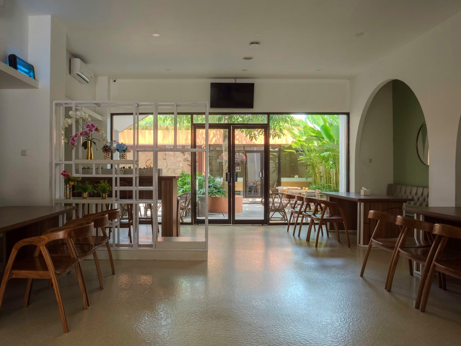 Bright empty cafe with wooden chairs, an arched alcove and glass doors opening onto a leafy courtyard