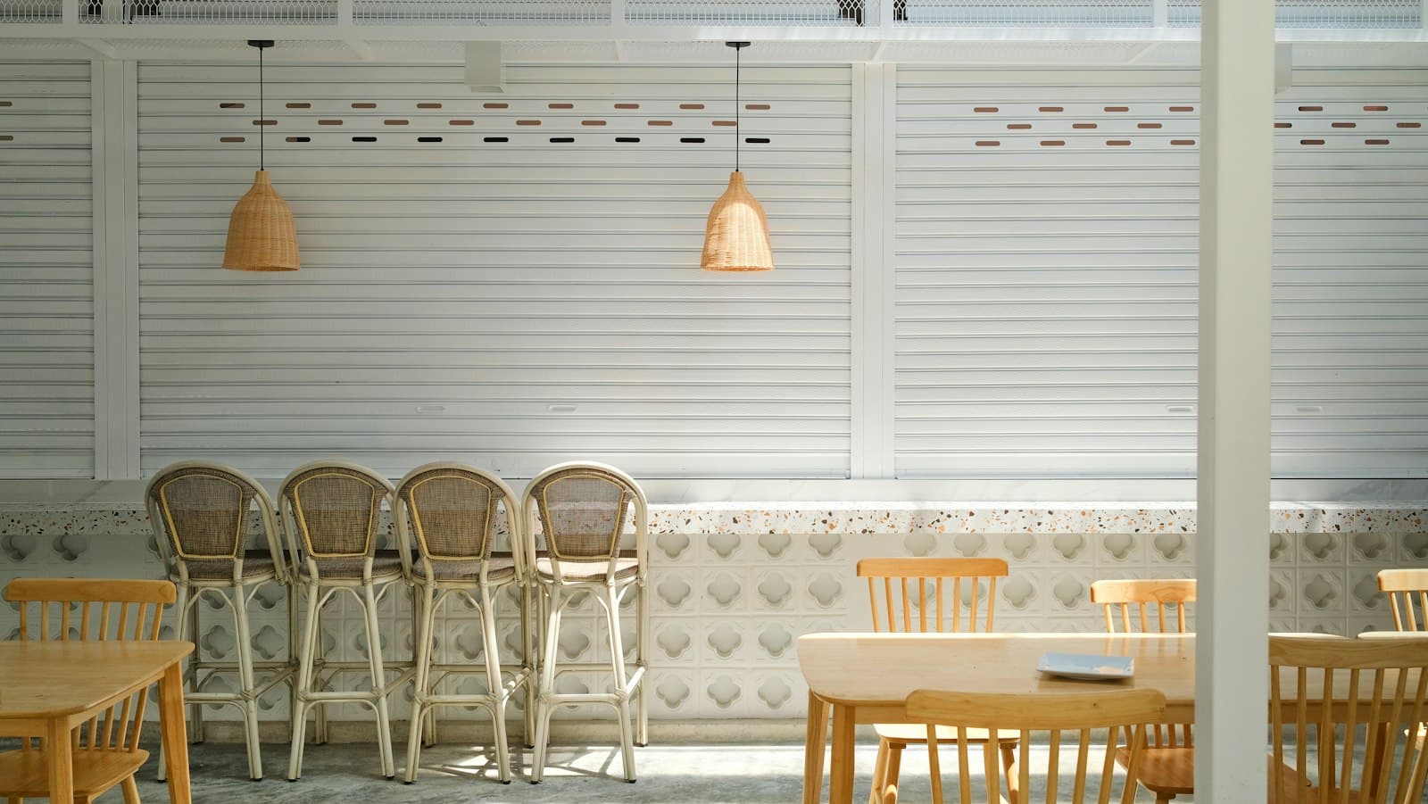 Sunlit cafe corner with rattan bar stools, pale wood tables and pendant lamps against white louvred walls