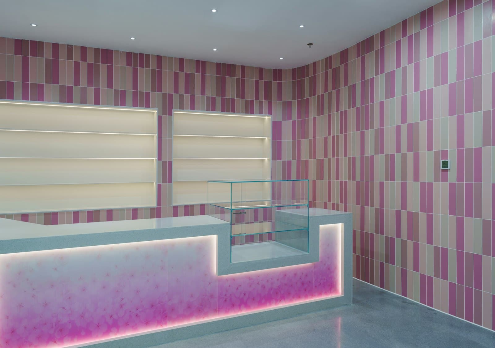 Empty retail counter with a glass display case and backlit shelving against pink and beige tiled walls