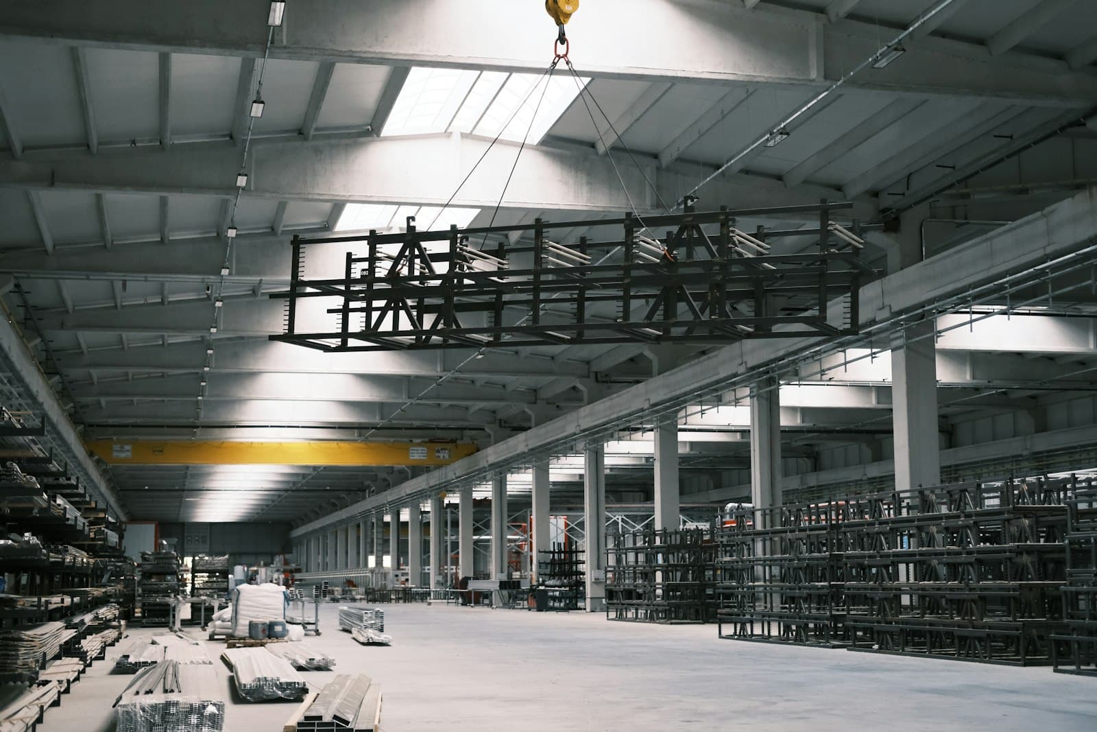 Bright factory bay where an overhead crane lifts a steel frame above racks of metal profiles and stacked stock