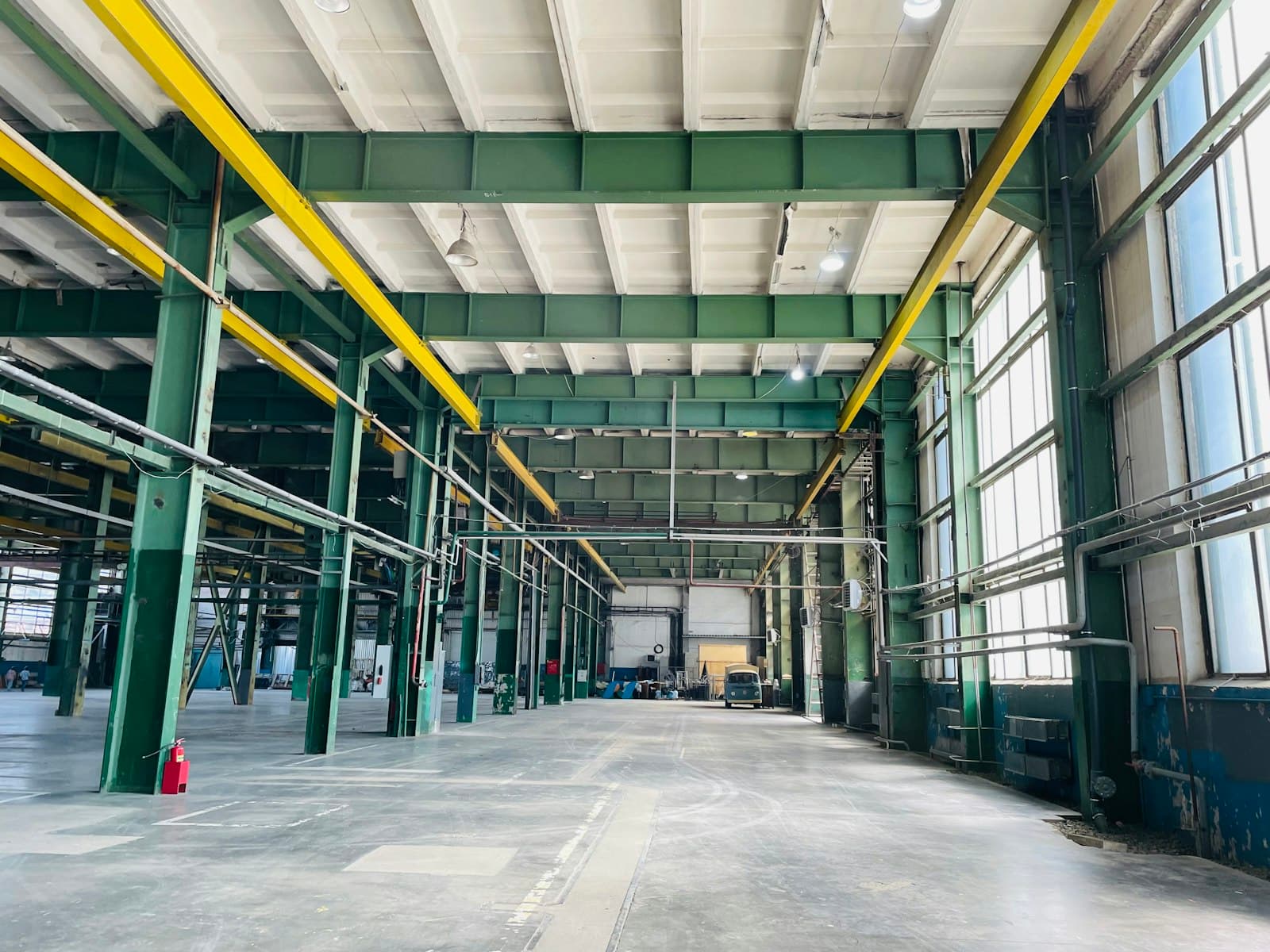 Empty factory floor with green steel columns and beams, yellow crane rails overhead and tall windows along one side