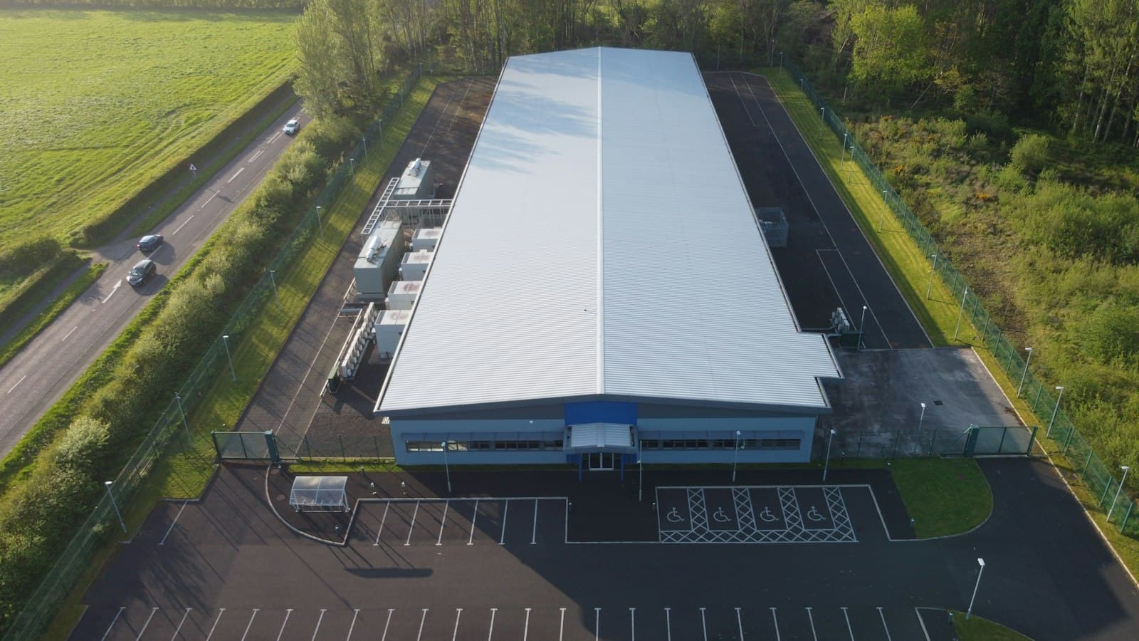Aerial view of a long industrial building with a pale metal roof, fenced perimeter, service yard and car park