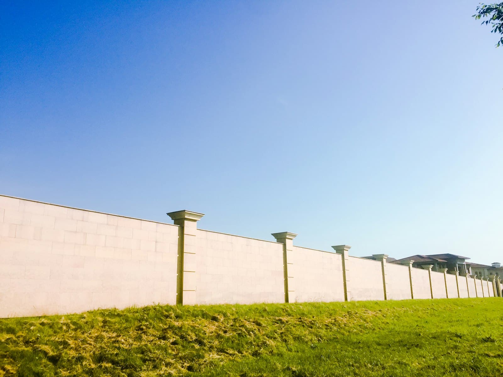 Long cream compound wall with square pillars running along a mown grass verge under a clear blue sky