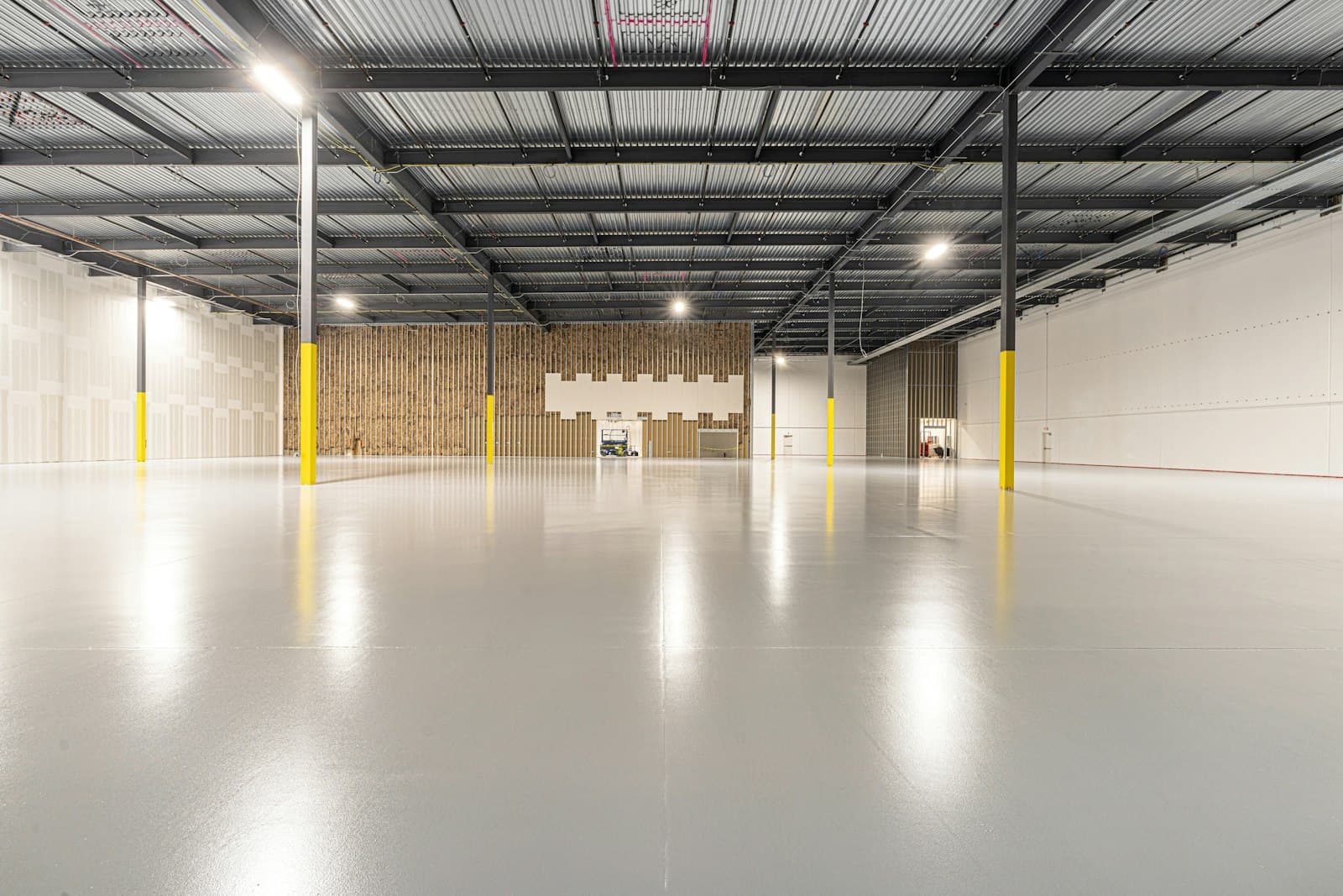 Empty high-bay warehouse with a polished grey floor, exposed steel roof deck, bright lights and yellow-guarded columns