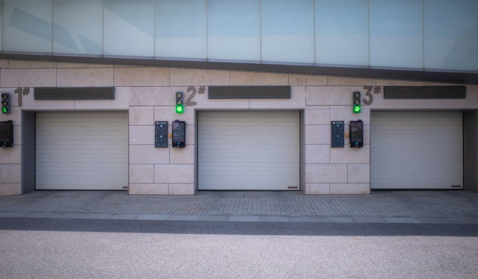 Three numbered white sectional loading bay doors with green signal lights and control panels in a pale stone-clad wall