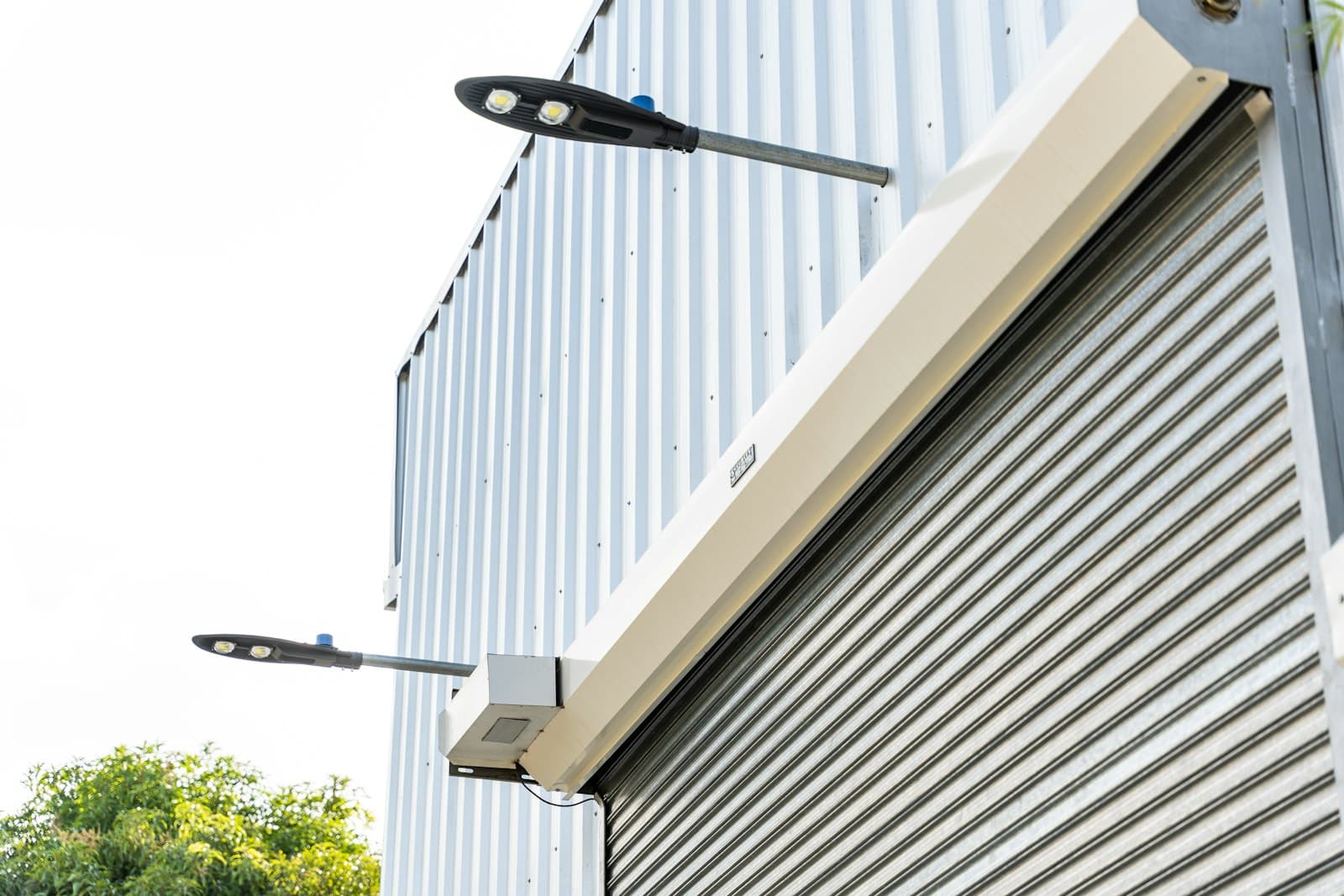 Close-up of a galvanised roller shutter and its motor housing under two LED floodlights on a metal-clad wall
