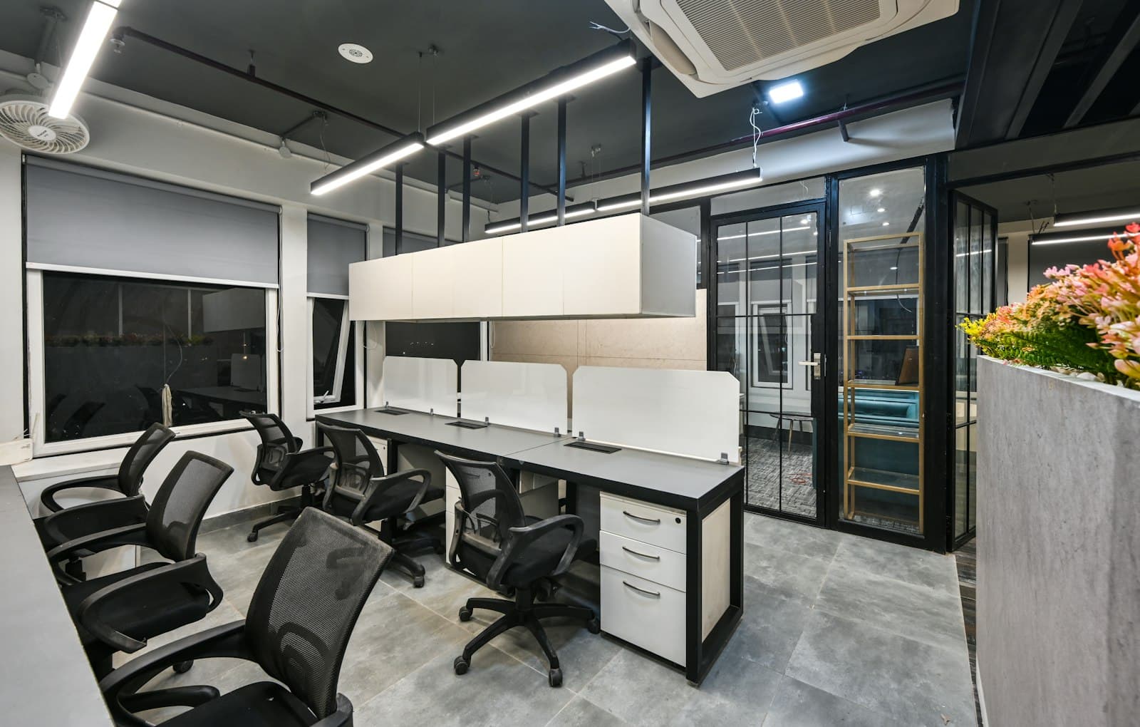 Compact office suite with a row of grey desks, white screens, mesh chairs and a black-framed glass cabin door