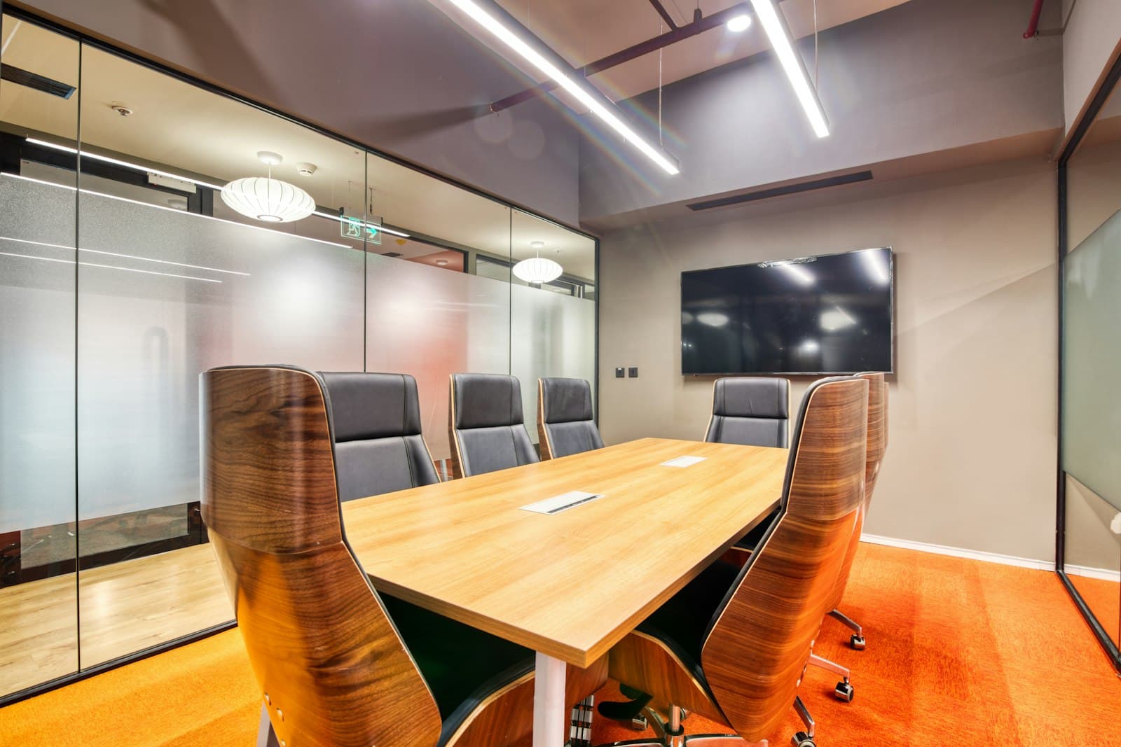 Small meeting room with frosted glass walls, a timber table, walnut-backed chairs, a wall screen and orange carpet