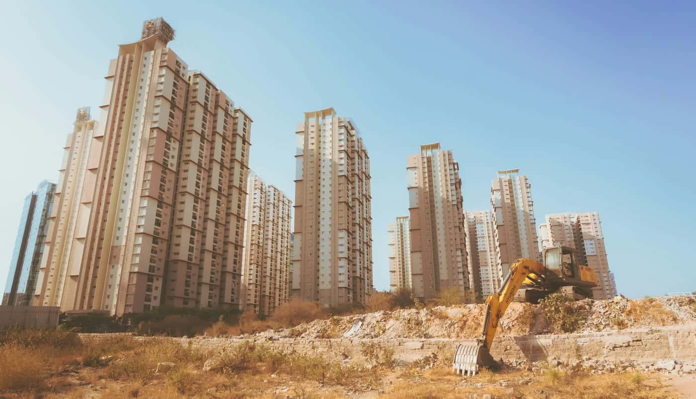 Cleared, levelled land with an excavator at work in front of a row of high-rise towers under a clear blue sky