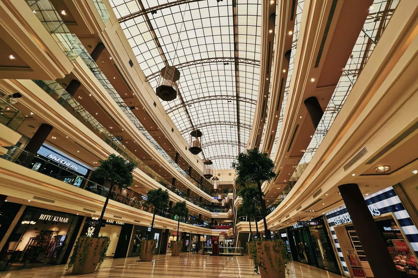 Three-level shopping mall atrium under a glazed barrel-vaulted roof, with shopfronts along each level and potted trees on a polished floor